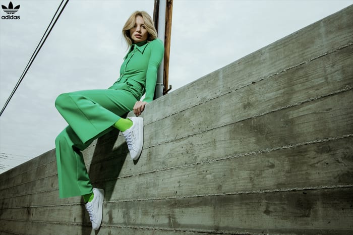 A fair-skinned woman in a green outfit sits on a concrete structure, wearing white sneakers and green socks, with the Adidas logo visible.