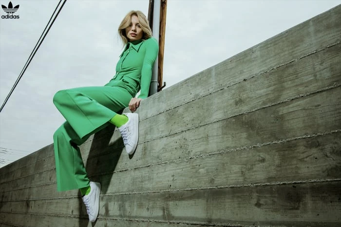 A fair-skinned woman in a green outfit sits on a concrete structure, wearing white sneakers and green socks, with the Adidas logo visible.