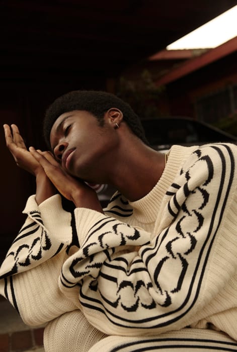 Young African American man in a patterned sweater leans his head on his hands outdoors with a dark background and dim lighting.