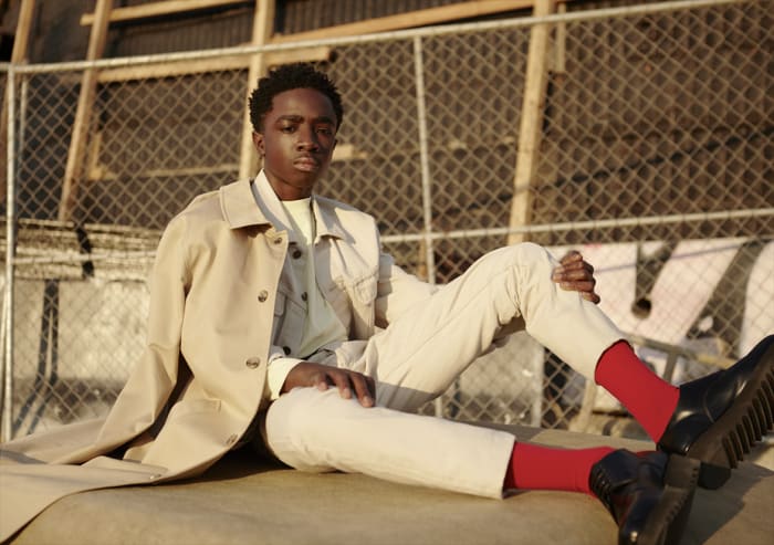 Young man in beige overcoat poses outdoors with bright red socks and black shoes, resting on hands with a chain-link fence background.
