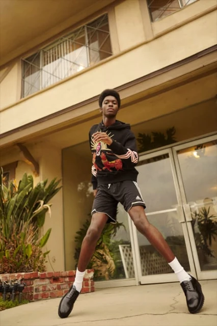 Young man in hoodie and athletic shorts poses outdoors in front of building with brick and foliage, wearing socks and dress shoes.