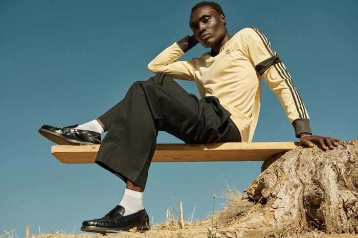 Dark-skinned adult man sitting on a wooden platform wearing an Adidas long sleeve shirt and black dress pants against a blue sky.