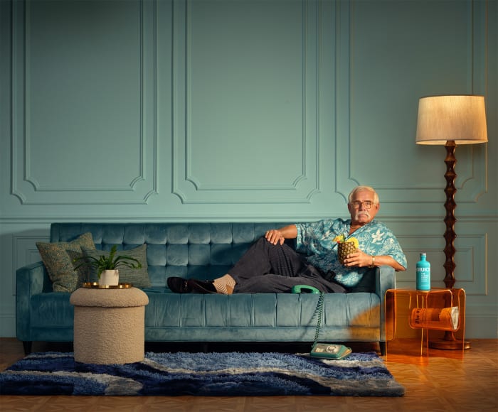Mature man lounges on a teal velvet couch with a pineapple cocktail in an opulent vintage-inspired interior, lit by a floor lamp.