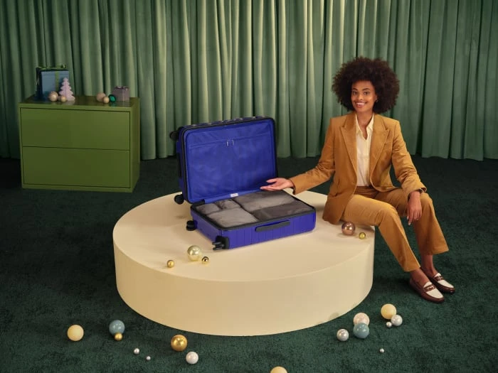 Stylish woman in a tan suit poses with an open, blue suitcase, surrounded by festive ornaments in a studio setting.