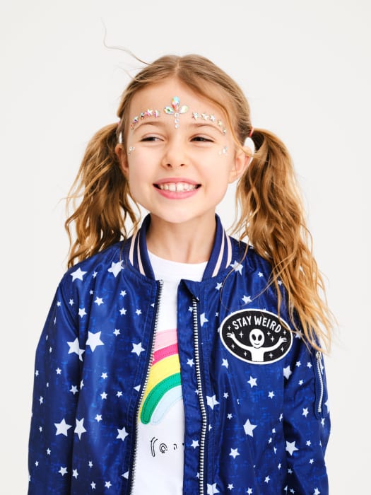 Smiling girl with glitter face decoration wears a blue star-patterned jacket. Head and shoulders, studio shot on a white backdrop.