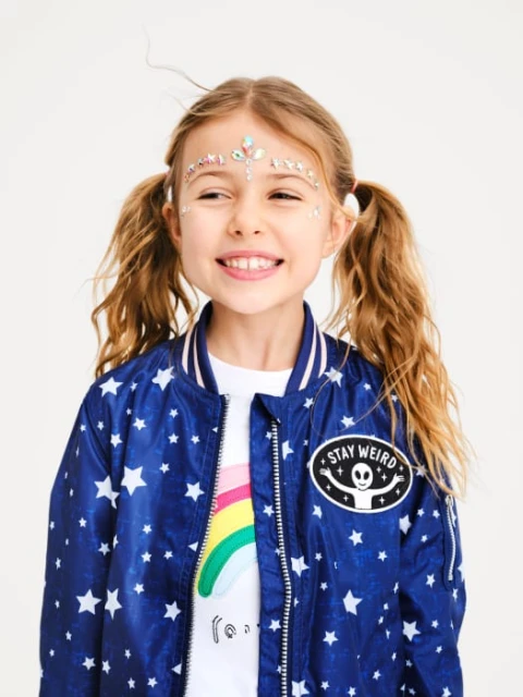 Smiling girl with glitter face decoration wears a blue star-patterned jacket. Head and shoulders, studio shot on a white backdrop.