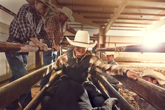 Cowboy riding bull in rodeo ring, captured indoors with spot lighting and multiple support people. Action shot with animal. 