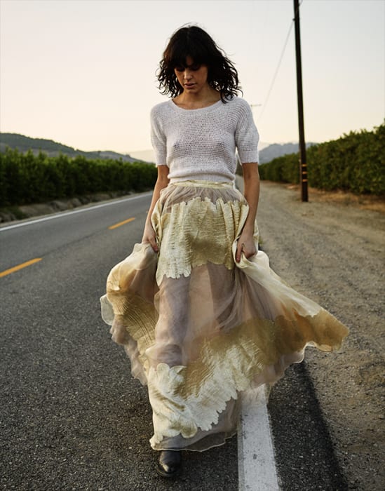 Adult woman in knit top and maxi skirt walking on an empty road flanked by rows of green foliage, trees, and utility poles.