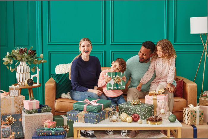 A happy multiracial family of four sits on a couch in front of a pile of wrapped Christmas gifts in an indoor setting with green walls.
