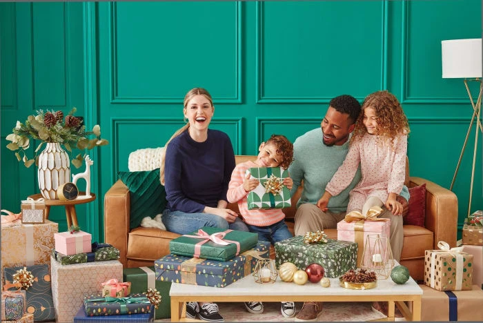 A happy multiracial family of four sits on a couch in front of a pile of wrapped Christmas gifts in an indoor setting with green walls.