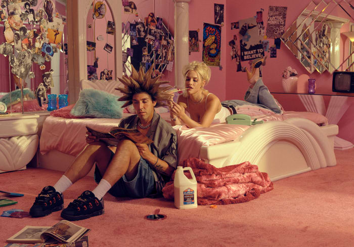 Two people in a pink bedroom: a male sitting on the pink carpet reading a magazine and a female lying on a bed with a cigarette.