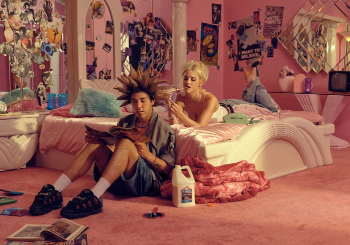 Two people in a pink bedroom: a male sitting on the pink carpet reading a magazine and a female lying on a bed with a cigarette.