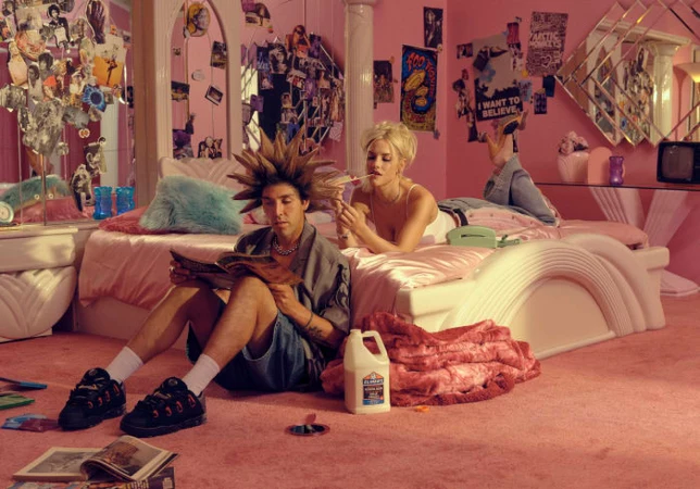 Two people in a pink bedroom: a male sitting on the pink carpet reading a magazine and a female lying on a bed with a cigarette.