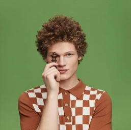 A man with curly red hair holding a wooden chess piece in front of his eye against a green background