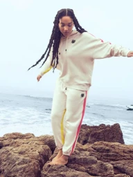 Woman in cream hoodie and sweatpants standing barefoot on rocky coastal shore