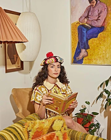 Woman wearing a floral crochet top and matching beret, holding a book in a vintage living room.