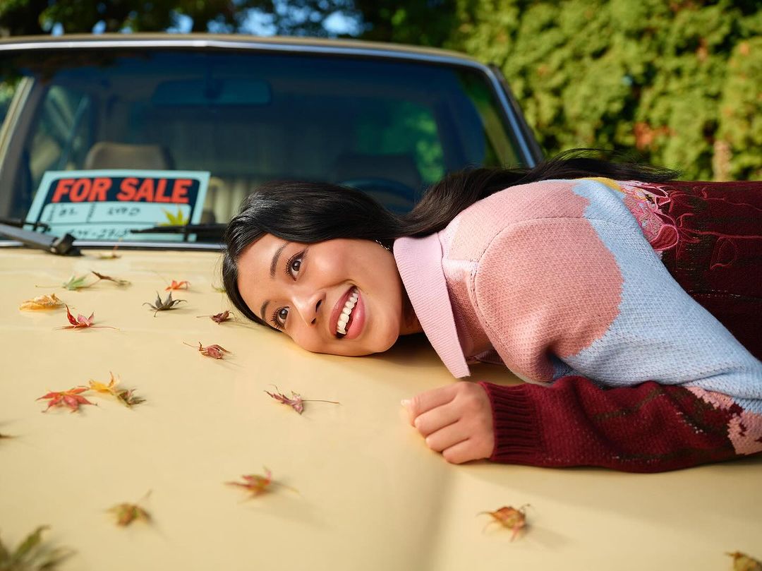 Smiling woman lying on the hood of a vintage car for sale, surrounded by fallen autumn leaves. Sunny, outdoor setting, medium shot.