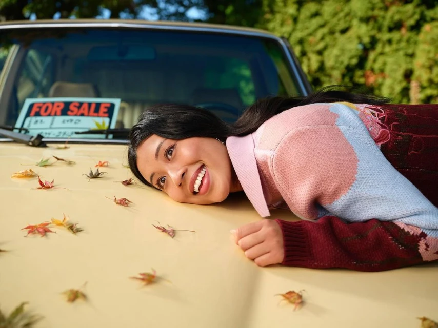 Smiling woman lying on the hood of a vintage car for sale, surrounded by fallen autumn leaves. Sunny, outdoor setting, medium shot.