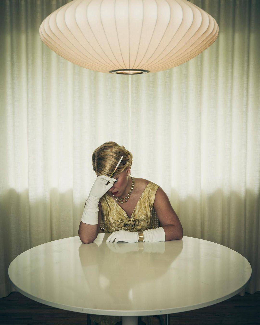 Blonde woman in gold dress sits at a round table with head in hand under a round mid-century modern light fixture and cream drapes.