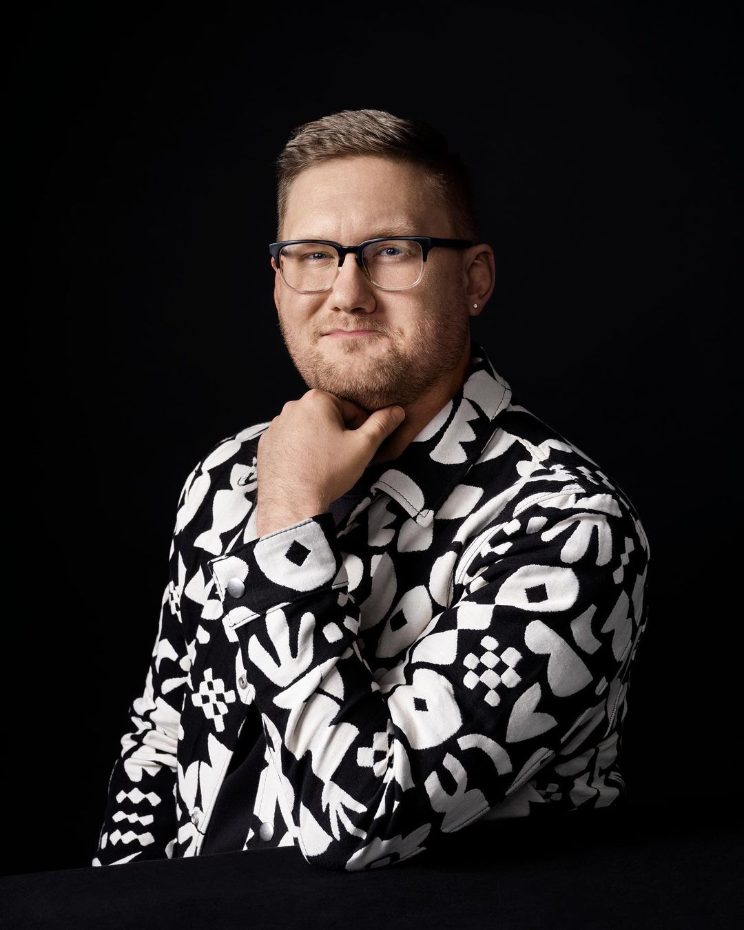 Portrait of an adult male with glasses, fair skin, and a beard, wearing a black-and-white patterned jacket against a dark background.