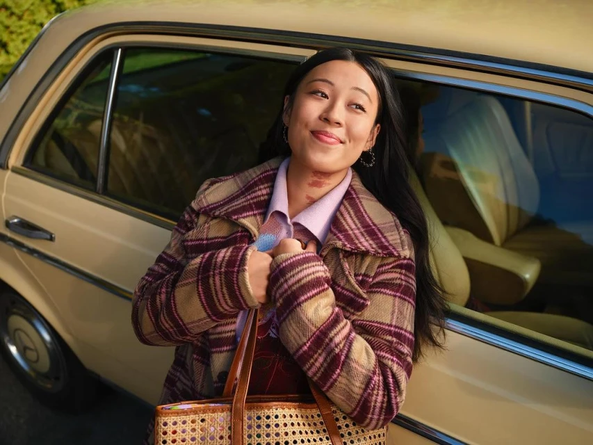 Smiling woman in a plaid coat stands next to a vintage car, holding a woven handbag. The scene is brightly lit and inviting.