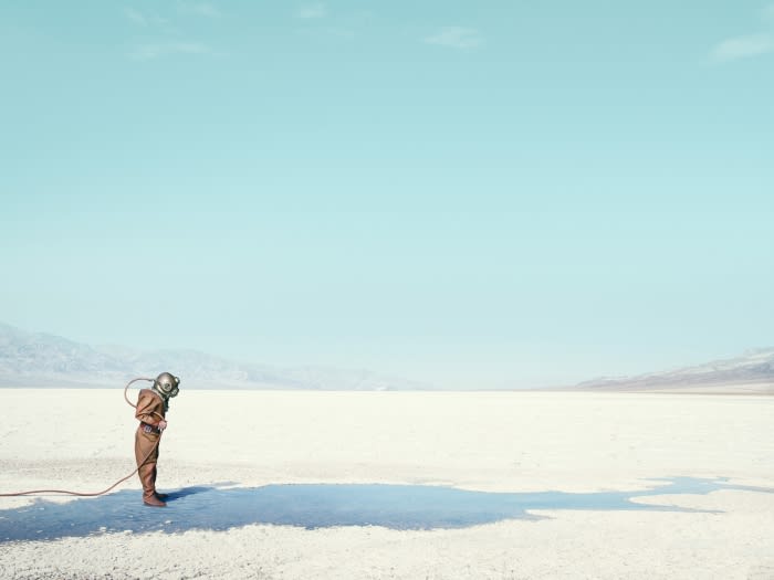 A lone person in vintage diving suit stands on a cracked, white desert floor under a bright blue sky in a conceptual fashion shot.