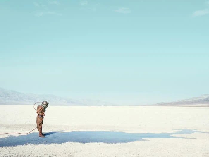 A lone person in vintage diving suit stands on a cracked, white desert floor under a bright blue sky in a conceptual fashion shot.