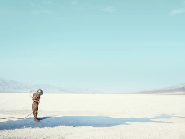 A lone person in vintage diving suit stands on a cracked, white desert floor under a bright blue sky in a conceptual fashion shot.