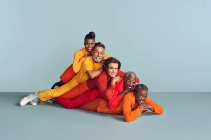 Group of five diverse people in orange and red outfits stacked playfully in a human pyramid against a grey studio backdrop.