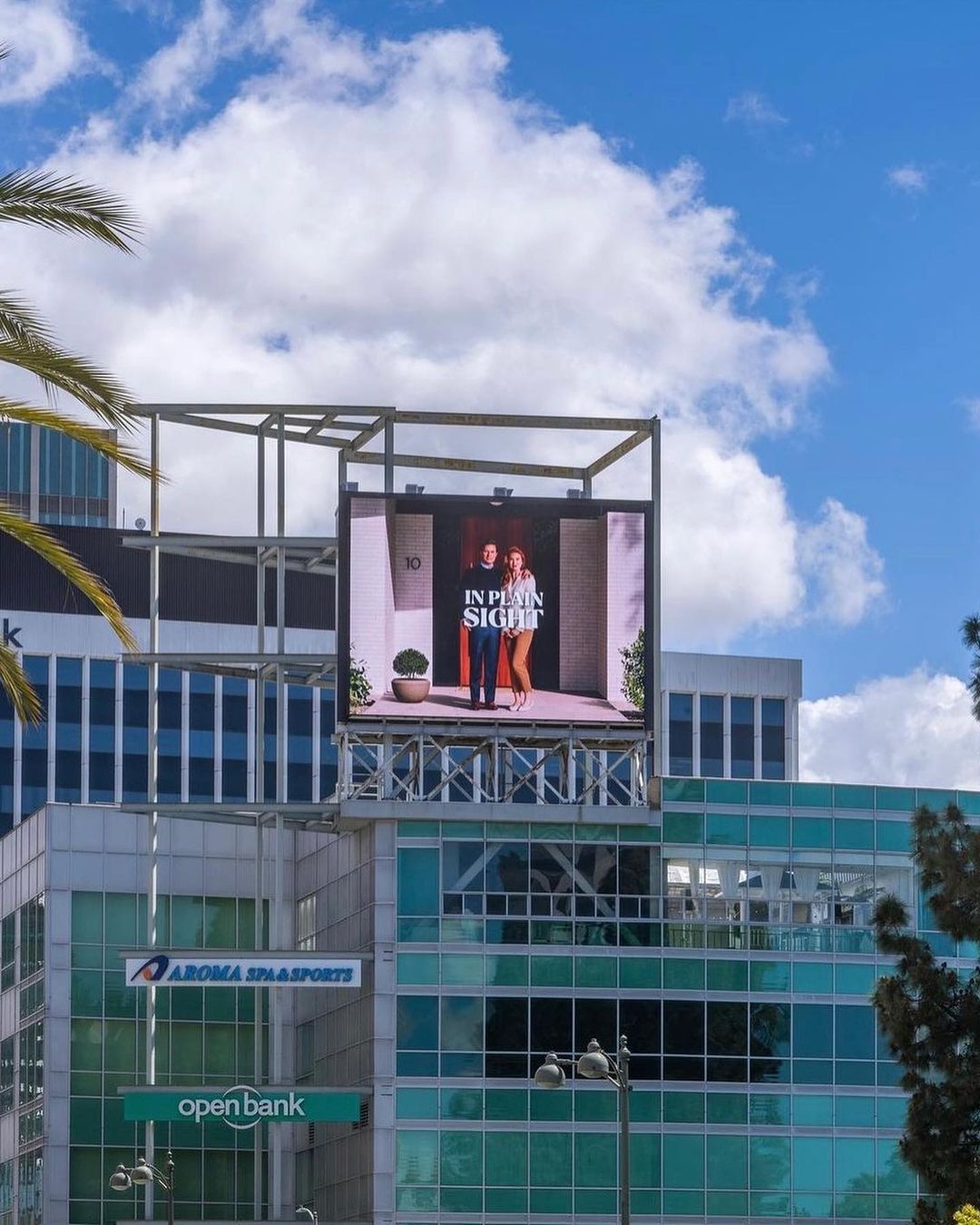 Outdoor advertising billboard displays two models standing at a doorway, promoting 'In Plain Sight' against a partly cloudy sky.