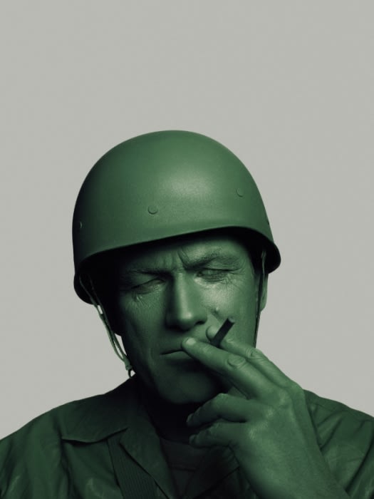 Close up of an adult man with green paint on his skin, wearing a toy soldier helmet, and holding a cigarette between his fingers.