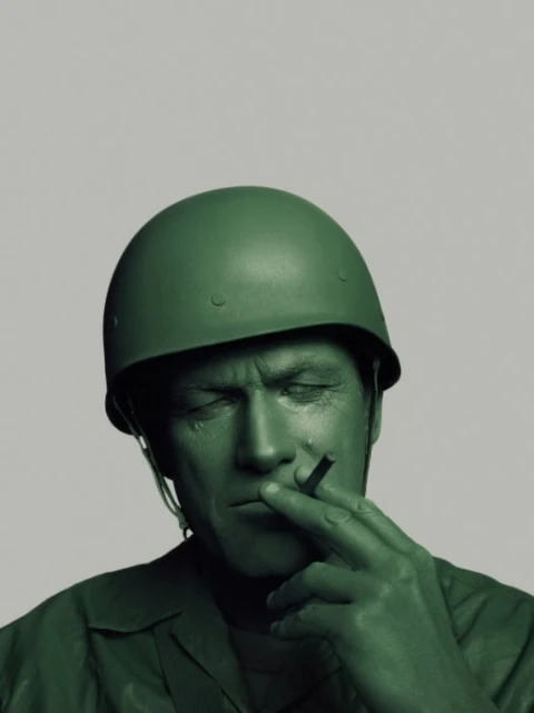 Close up of an adult man with green paint on his skin, wearing a toy soldier helmet, and holding a cigarette between his fingers.
