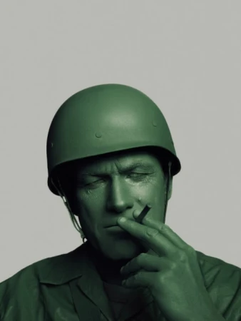 Close up of an adult man with green paint on his skin, wearing a toy soldier helmet, and holding a cigarette between his fingers.
