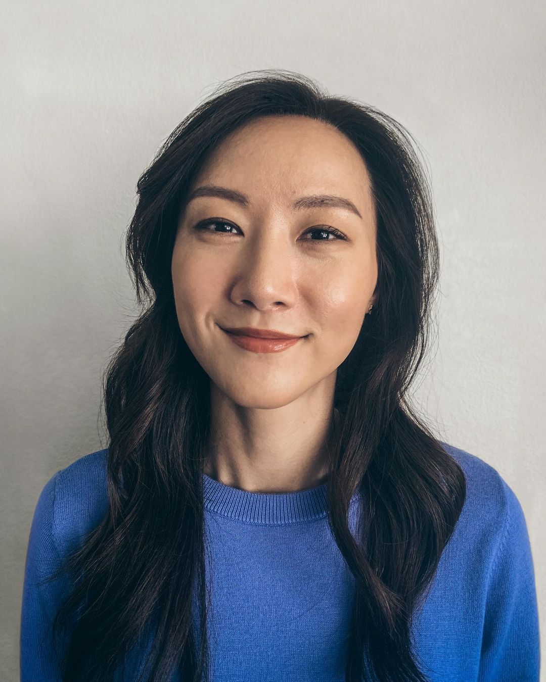 Portrait of a smiling Asian woman with long dark hair, wearing a blue sweater, against a light gray wall. Head and shoulders shot.
