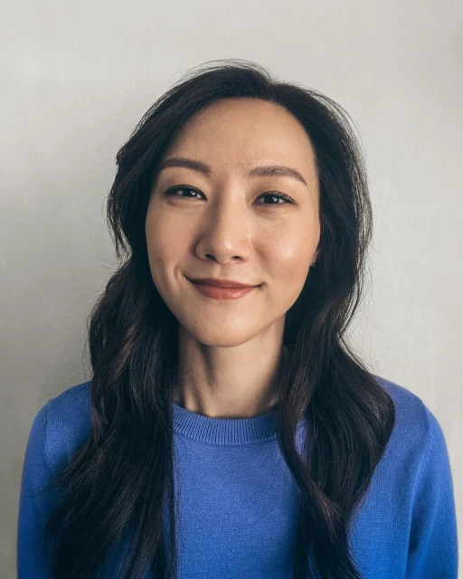 Portrait of a smiling Asian woman with long dark hair, wearing a blue sweater, against a light gray wall. Head and shoulders shot.