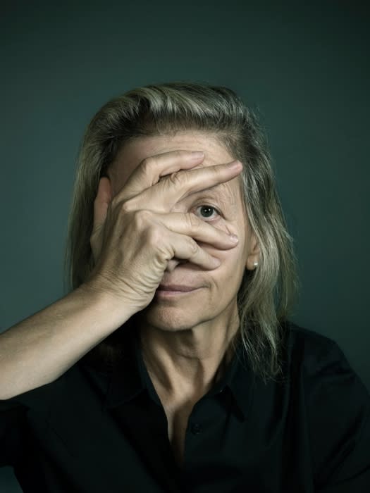 Portrait of a mature woman with light hair covering half her face with her hand against a dark-green background in studio lighting.