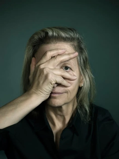 Portrait of a mature woman with light hair covering half her face with her hand against a dark-green background in studio lighting.