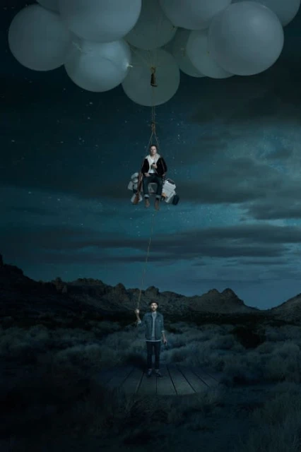 Creative composite of a man floating in a balloon chair, held by another man on a wooden dock in a desert landscape under a starry sky.