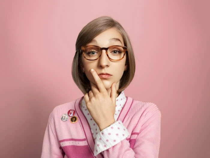 Young woman with glasses in pink cardigan, resting finger on chin against a pink background, creating a contemplative and stylish portrait.