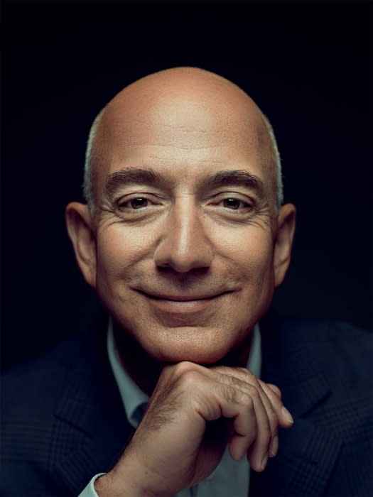 Portrait of a bald adult male in a dark suit and light blue shirt, resting his chin on his hand against a dark background.