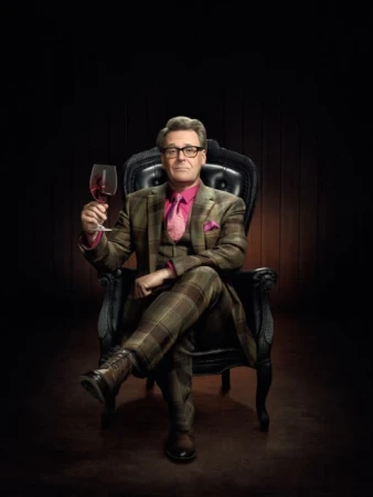 Mature man in glasses and plaid suit sits in a black throne-like chair holding a glass of red wine against a dark background.