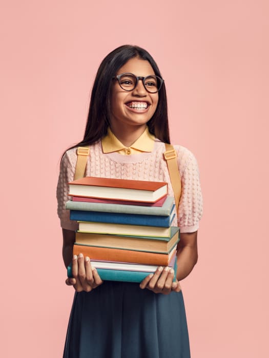 Smiling woman holding a stack of books. She is wearing glasses and a yellow shirt with a pink knit sweater and a blue skirt.