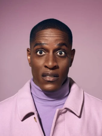 Close-up shot of a Black man with a surprised expression, wearing a lavender turtleneck and a light pink overcoat, against a pink background.