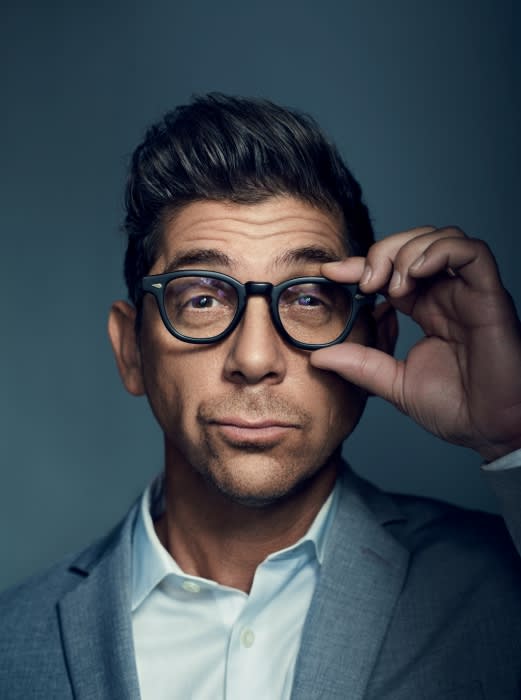 Portrait of adult male in a blazer, shirt and eyeglasses, adjusting his glasses with his right hand against a blue-grey background.