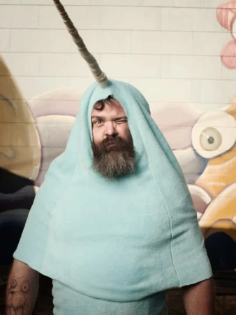 Man with beard wearing a light blue narwhal costume headpiece with a tusk, standing in front of a painted mural wall.