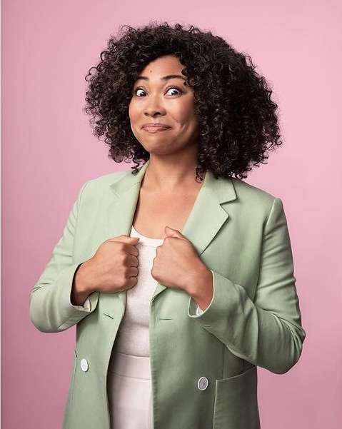 Portrait of a woman with curly hair wearing a blazer against a pink background. She has a funny expression, and her hands are at her chest.