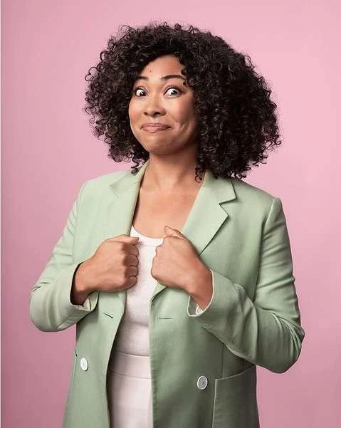 Portrait of a woman with curly hair wearing a blazer against a pink background. She has a funny expression, and her hands are at her chest.