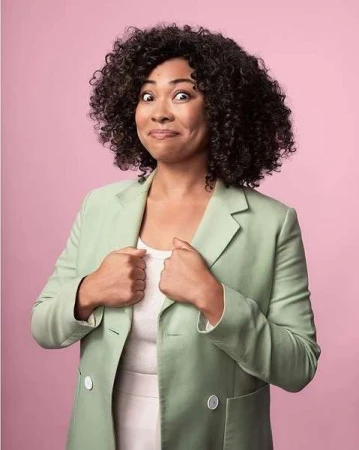 Portrait of a woman with curly hair wearing a blazer against a pink background. She has a funny expression, and her hands are at her chest.