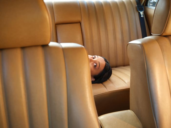 Woman with dark hair peeks between tan leather car seats in an indoor setting, creating a somewhat concealed and intriguing composition.