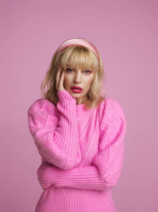 Blonde woman with a pink headband poses in front of a pink backdrop, wearing a pink sweater and lipstick. Portrait-style shot.